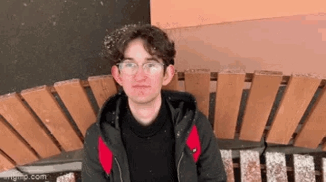 a young man wearing glasses is sitting on a wooden bench in the snow .