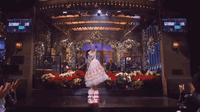 a woman in a white dress stands on a stage in front of a saturday night live screen