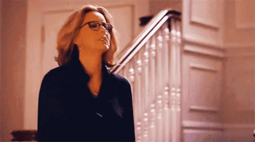 a woman wearing glasses and a black coat is standing in front of a staircase .