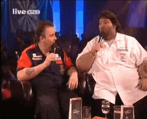 two men are sitting at a table and one is holding a microphone while the other talks into it ..