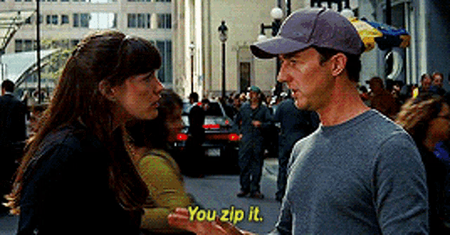 a man in a baseball cap is talking to a woman in a crowded street and the woman is saying you zip it