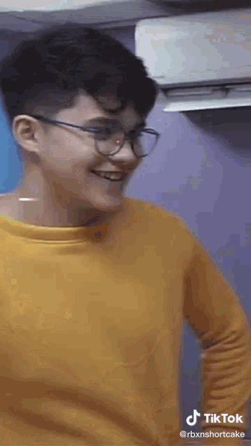 a young man wearing glasses and a yellow shirt is smiling and standing in front of an air conditioner .