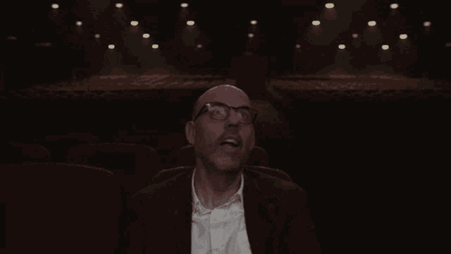 a bald man wearing glasses is sitting in an auditorium and pointing up