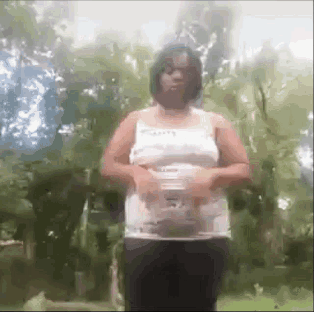 a woman in a white tank top and black pants is standing in front of a forest .