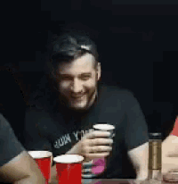 a man in a black shirt is sitting at a table holding a cup of beer .