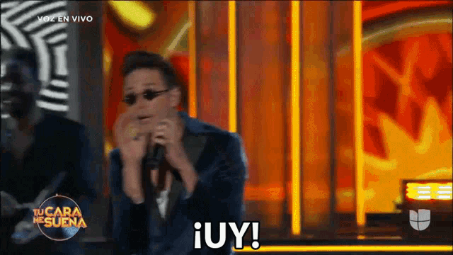 a man in a suit is singing into a microphone while wearing sunglasses and saying ¡ uy !