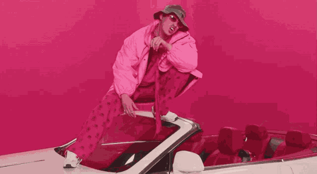 a man in a pink jacket is squatting on the hood of a white convertible car