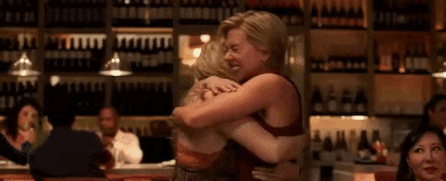 two women are hugging each other in a bar in front of a wall of wine bottles .