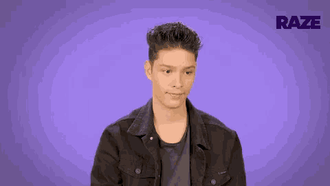 a young man in a black jacket is standing in front of a purple background and making a funny face .