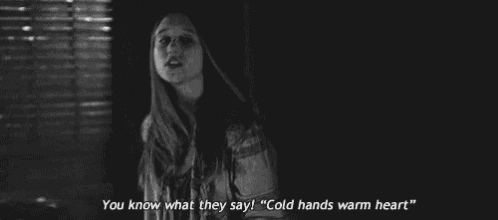 a black and white photo of a woman standing in a dark room and saying `` cold hands warm heart '' .
