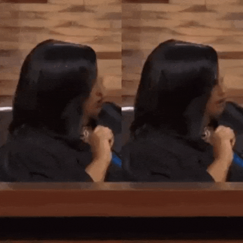 a woman with long black hair is sitting at a table with her hands on her chest .