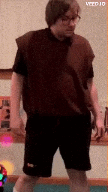 a man in a brown vest and black shorts is standing in front of a wall with the words veed.io below him