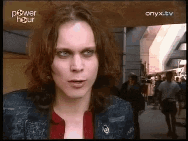 a man with long hair and blue eyes is standing in front of a crowd of people on a street .
