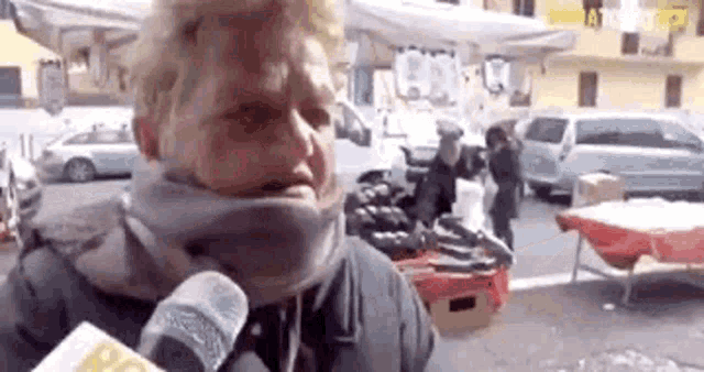 a woman wearing a scarf is talking into a microphone while standing in front of a market .