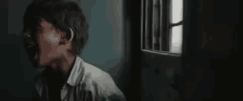 a man in a white shirt is standing in a dark room looking out a window .
