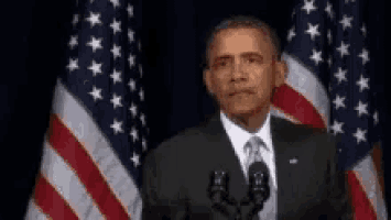 barack obama is giving a speech in front of a microphone while standing in front of american flags .