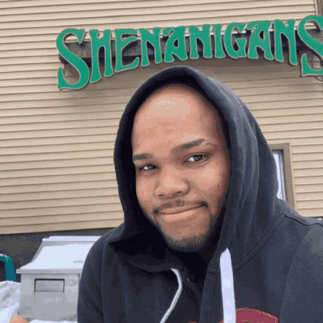 a man wearing a black hoodie stands in front of a green sign that says shenanigans
