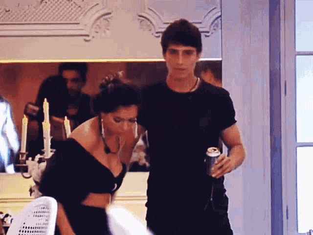 a man in a black shirt is holding a can of beer while standing next to a woman in a black crop top