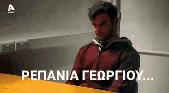 a man in a red jacket sits at a table in front of a sign that says pepania georgiou