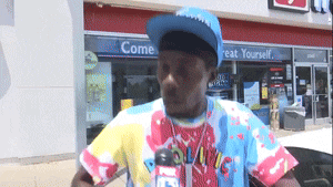 a man wearing a tie dye shirt stands in front of a store that says come treat yourself