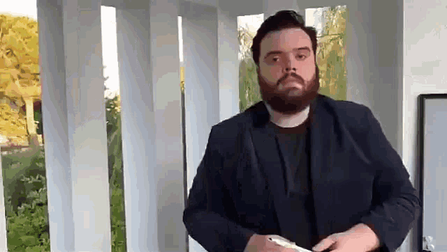 a man with a beard is standing in front of a white wall and holding a cell phone .