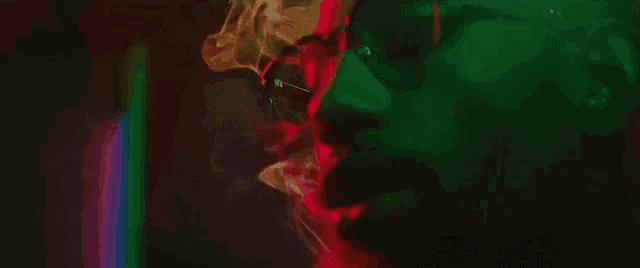 a man wearing glasses is smoking a cigarette in a dark room with red and green lights .