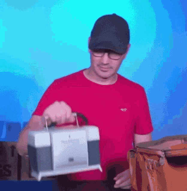 a man in a red shirt and black hat is holding a cooler with a handle .