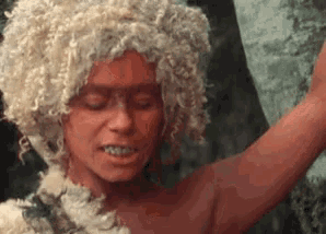 a woman with white curly hair and red skin is standing in front of a tree .