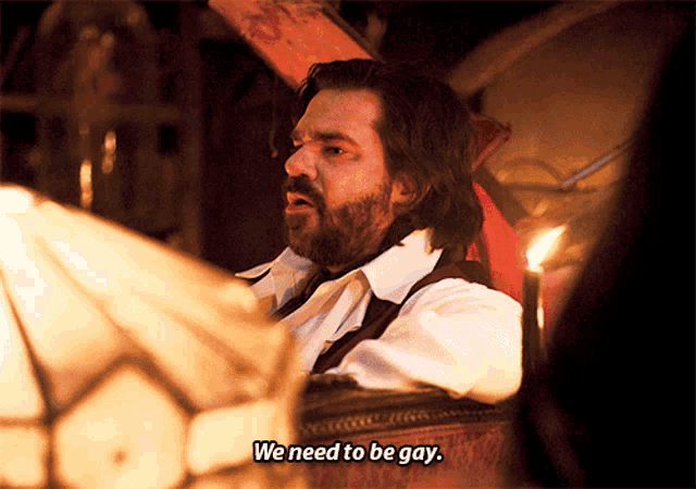 a man with a beard is sitting in a chair and says we need to be gay