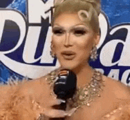 a drag queen is talking into a microphone while wearing a feathered dress .
