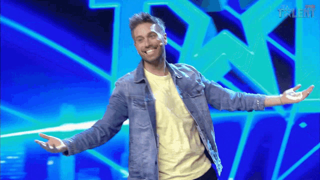 a man in a denim jacket stands on a stage with his arms outstretched in front of a screen that says talent