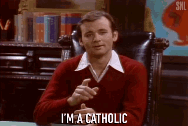 a man in a red sweater is sitting in a chair and saying i 'm a catholic .