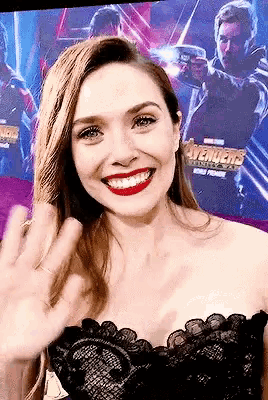 a woman is smiling and waving at the camera while wearing a black dress .