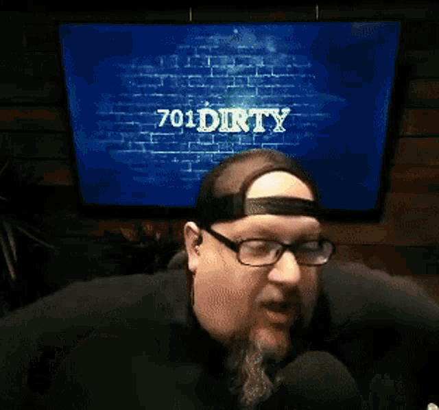 a man with a beard and glasses is talking into a microphone in front of a television screen that says tommy