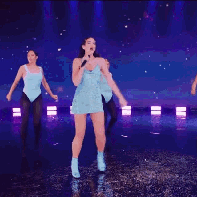 a woman in a light blue dress is singing into a microphone on a stage surrounded by dancers .