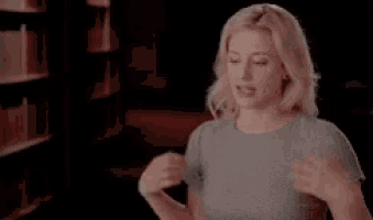 a woman in a grey shirt is standing in front of a bookshelf and talking to someone .