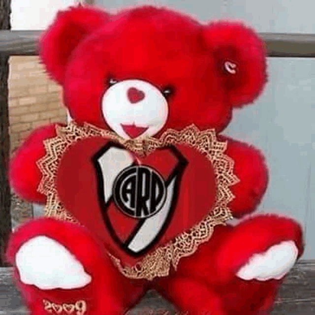 a red teddy bear is sitting on a bench holding a red heart with a logo on it .
