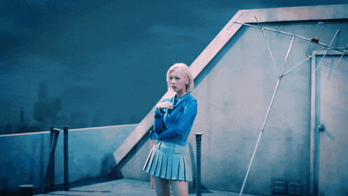 a woman in a blue jacket and pleated skirt stands on a rooftop