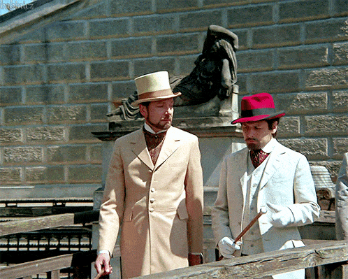 a man in a top hat stands next to another man in a white suit and white gloves