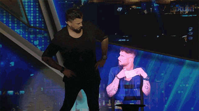 a man in a black shirt stands in front of a projection of a man in a blue shirt