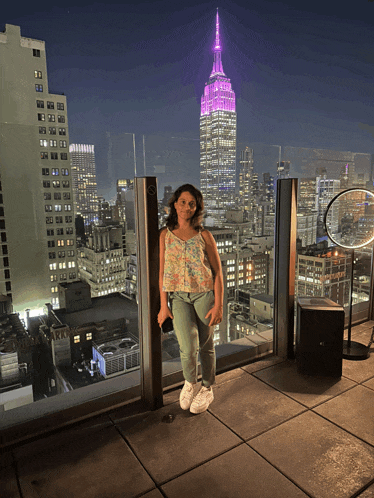 a woman stands on a balcony with the empire state building lit up in pink
