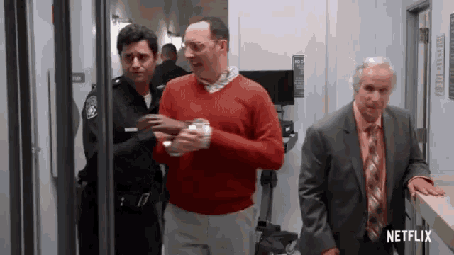 a man in a red sweater is being escorted by a police officer and a man in a suit and tie