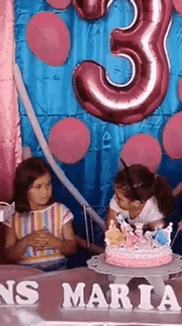 two little girls are sitting at a table with a cake and balloons in the background .
