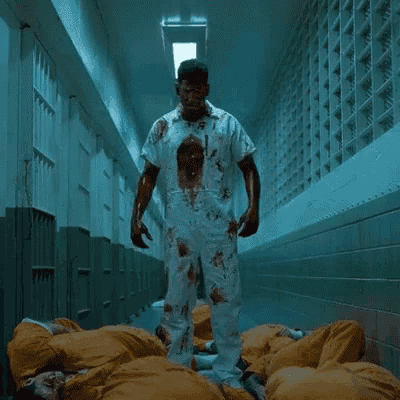 a man with blood on his shirt is standing in a hallway surrounded by prisoners in orange jumpsuits