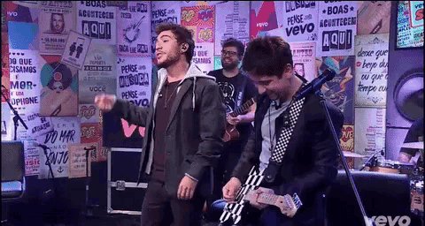 a man singing into a microphone while another man plays a guitar in front of a wall with posters that say aqui