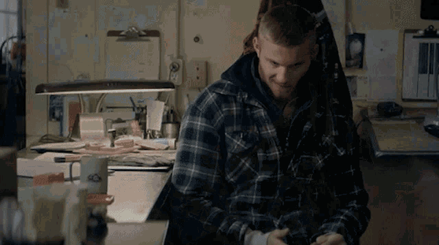 a man in a plaid shirt sits at a desk in front of a lamp