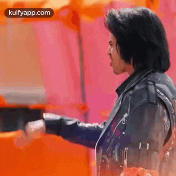 a man in a leather jacket is standing in front of a pink wall and holding something in his hand .