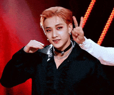 a young man with red hair is wearing a choker and a necklace and giving a peace sign .