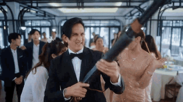 a man in a tuxedo is holding a gun in front of a group of people .