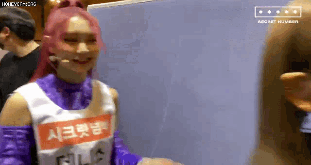 a woman with pink hair is wearing a purple and white outfit and smiling while standing in front of a white board .